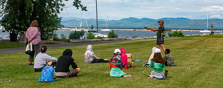 Group sitting on waterfront lawn watching instructor fake cast a rod during a fishing class at Perkins Pier. Lake Champlain and Adirondacks in the background