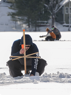 Person squatting to set tip-up in an ice fishing hole on a frozen lake, a couple out of focus also doing the same thing behind him